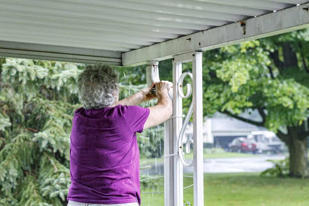 Drenched senior adult woman repairing overflowing roof downspout
